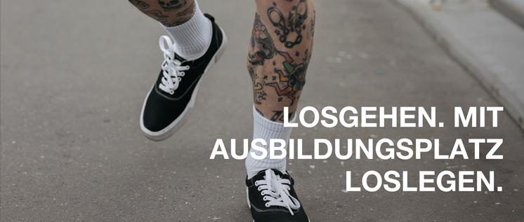 Tättoowierte Beine in schwarzen Sneakern und weißen Socken, die über eine Straße laufen.