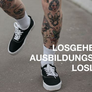 Tättoowierte Beine in schwarzen Sneakern und weißen Socken, die über eine Straße laufen.