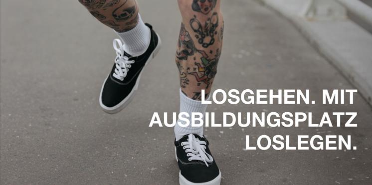 Tättoowierte Beine in schwarzen Sneakern und weißen Socken, die über eine Straße laufen.