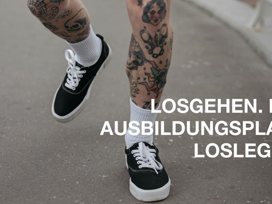 Tättoowierte Beine in schwarzen Sneakern und weißen Socken, die über eine Straße laufen.