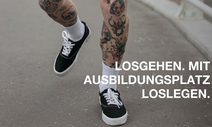 Tättoowierte Beine in schwarzen Sneakern und weißen Socken, die über eine Straße laufen.
