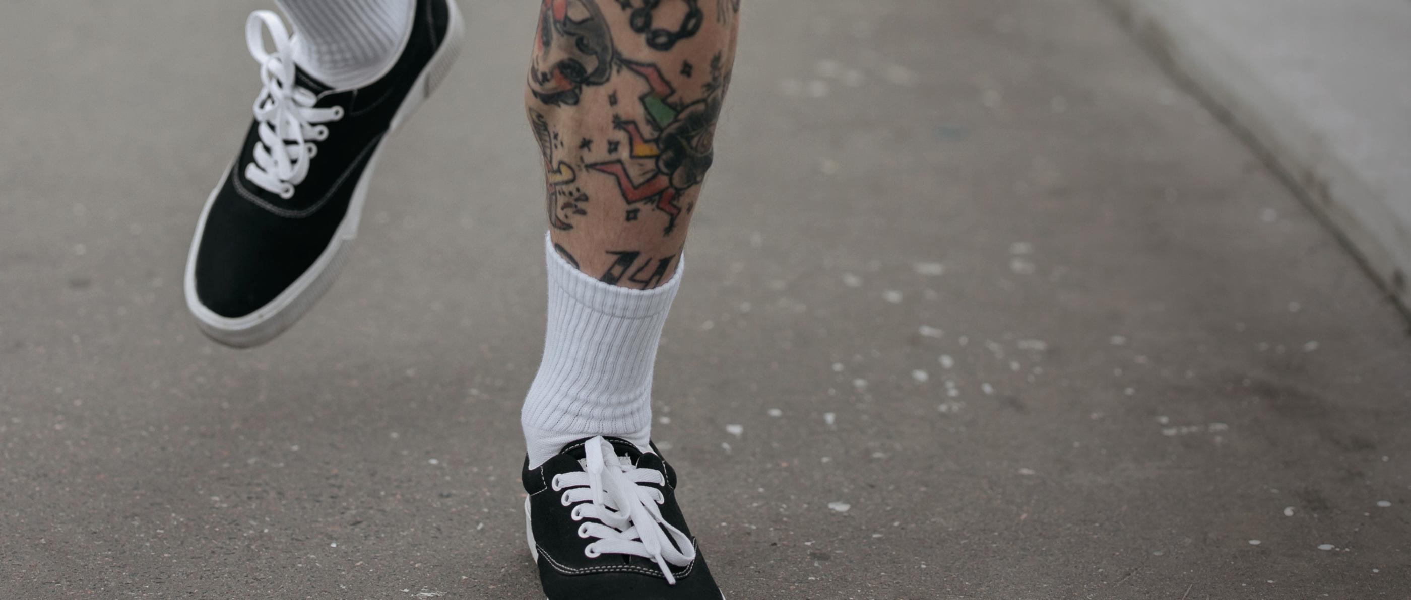 Tättoowierte Beine in schwarzen Sneakern und weißen Socken, die über eine Straße laufen.