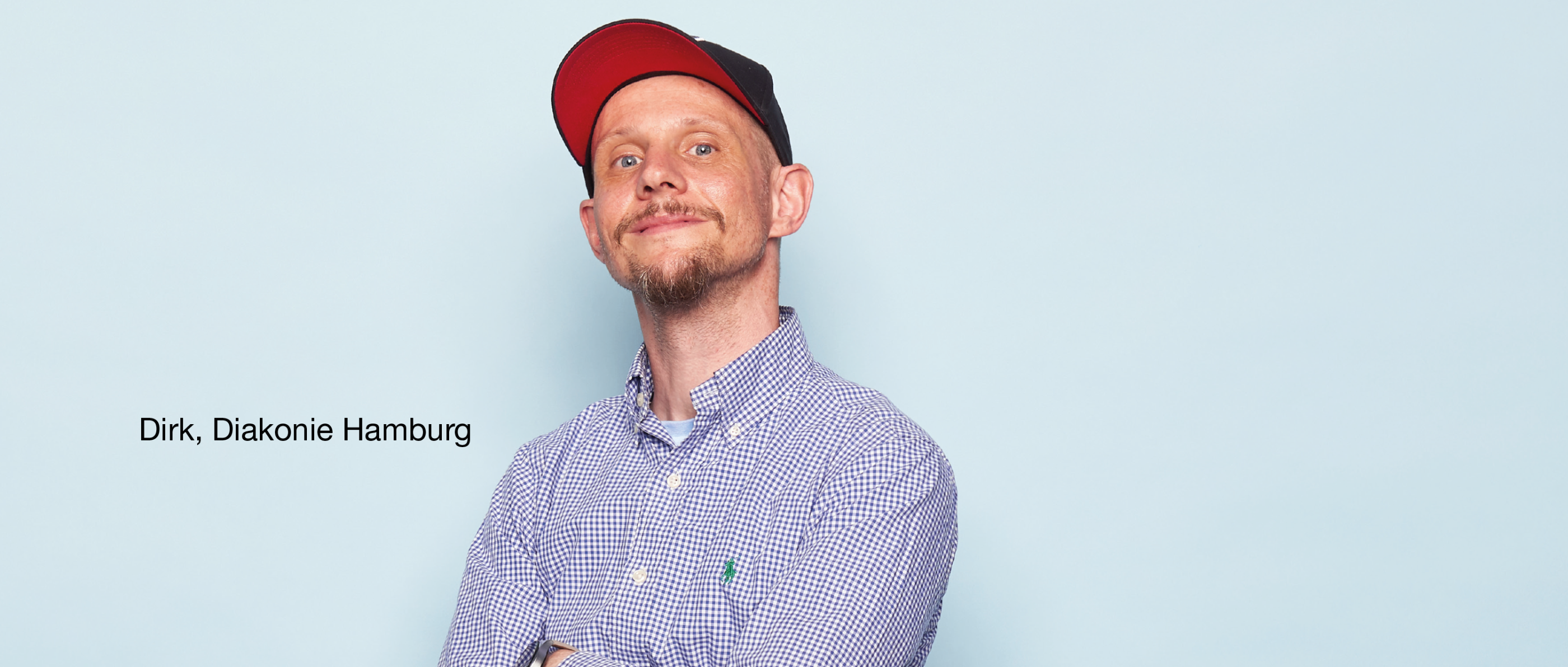 Person mit Bart, kariertem Hemd und schwarzer Mütze mit rotem Schirm steht mit verschränkten Armen vor hellblauem Hintergrund. Text: „Dirk, Diakonie Hamburg“.