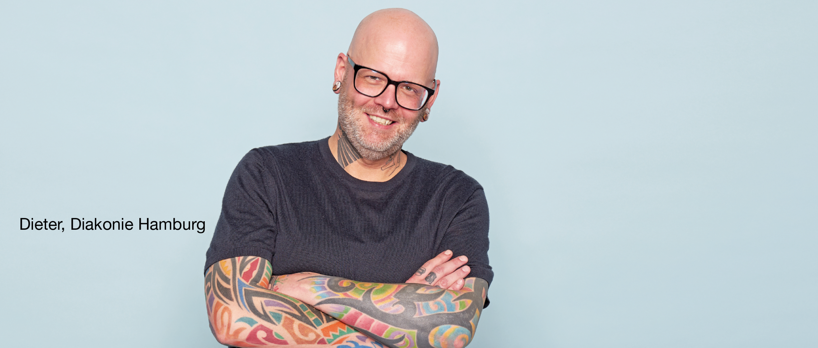 Person mit Brille und sichtbaren Tattoos trägt ein dunkles Shirt, lächelt und steht mit verschränkten Armen vor hellblauem Hintergrund. Text: „Dieter, Diakonie Hamburg“.