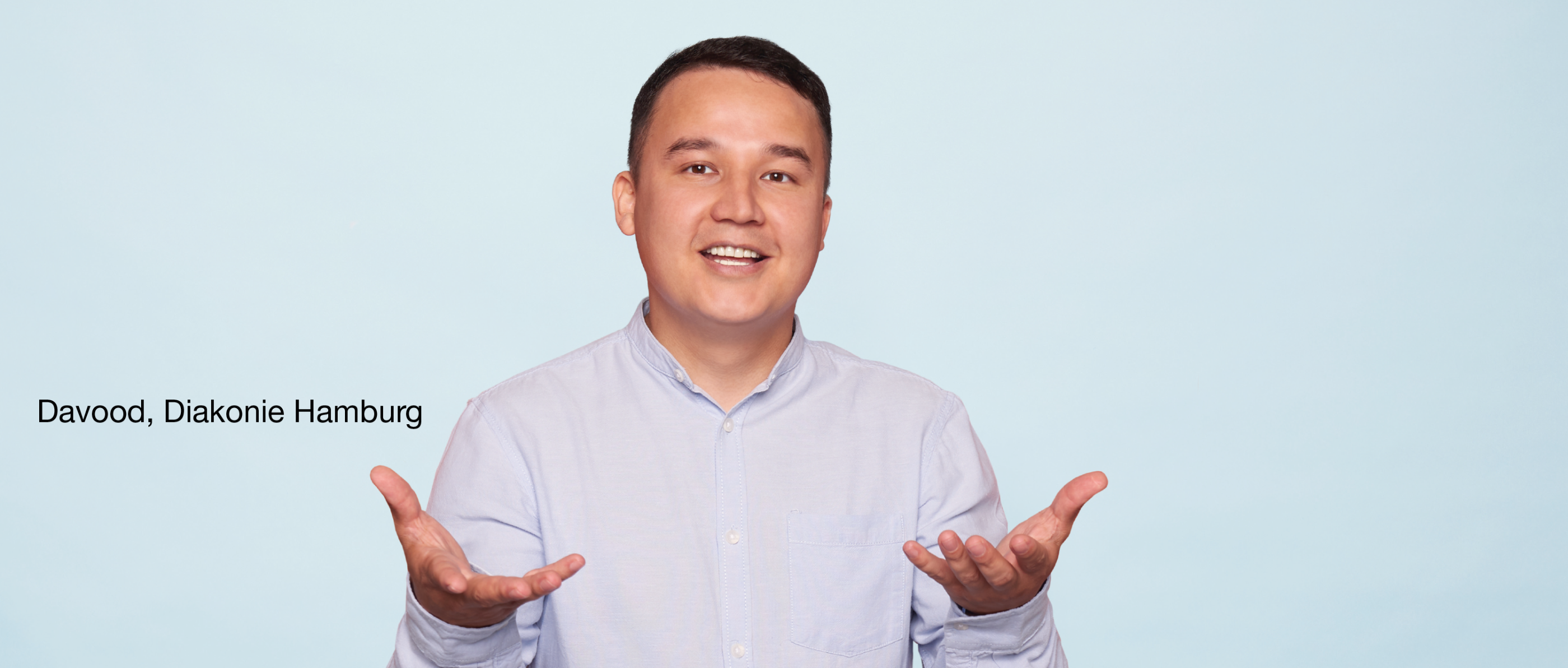 Person mit kurzen dunklen Haaren in hellblauem Hemd gestikuliert freundlich vor hellblauem Hintergrund. Text: „Davood, Diakonie Hamburg“.