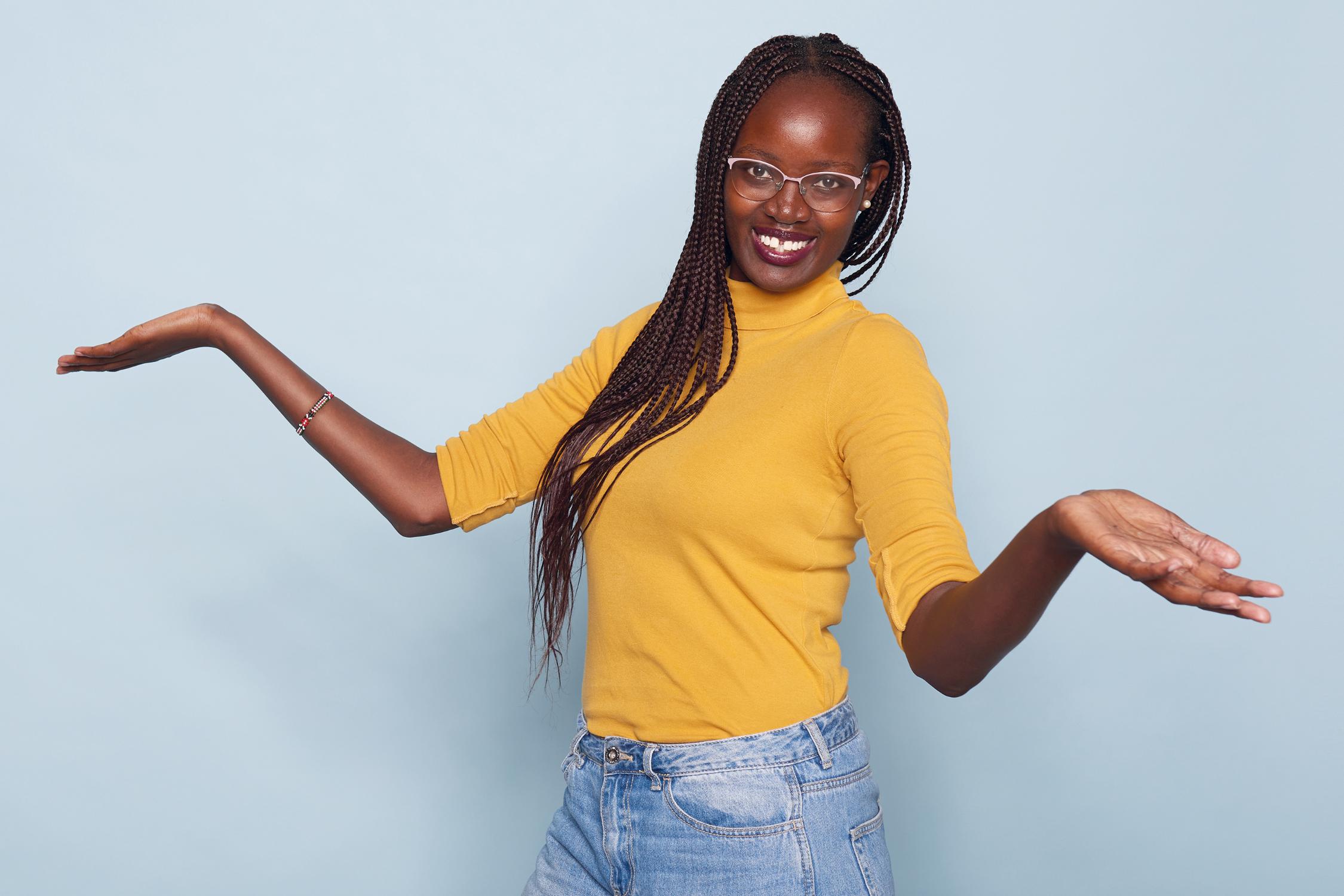 Person in gelbem Pulli mit ausgebreiteten Armen vor hellblauem Hintergrund.