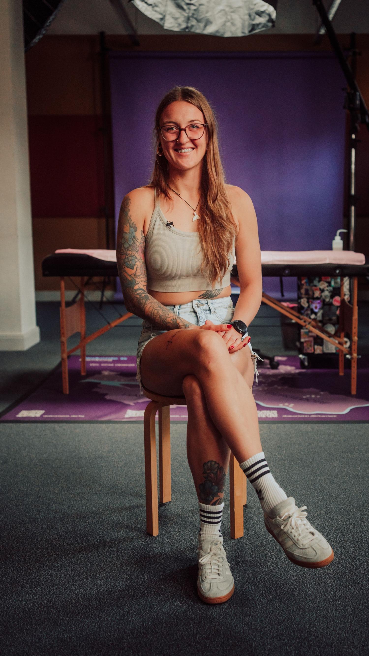 Person mit langen Haaren und Brille sitzt auf einem Hocker vor violettem Hintergrund. Trägt ein helles ärmelloses Oberteil, sichtbare Tattoos an einem Arm, Behandlungsliege im Hintergrund.