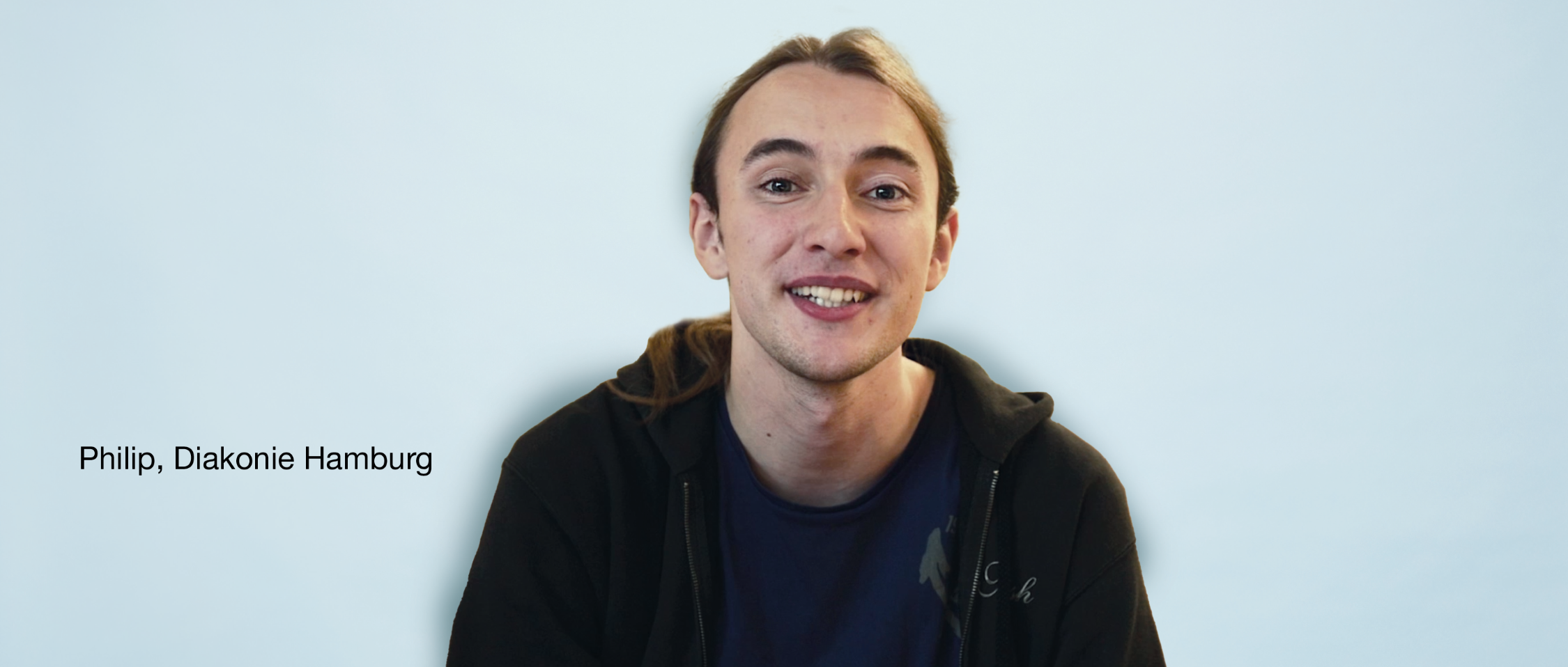 Person mit zurückgebundenem Haar im schwarzen Hoodie über dunklem Shirt lächelt vor hellblauem Hintergrund. Text links: „Philip, Diakonie Hamburg“.