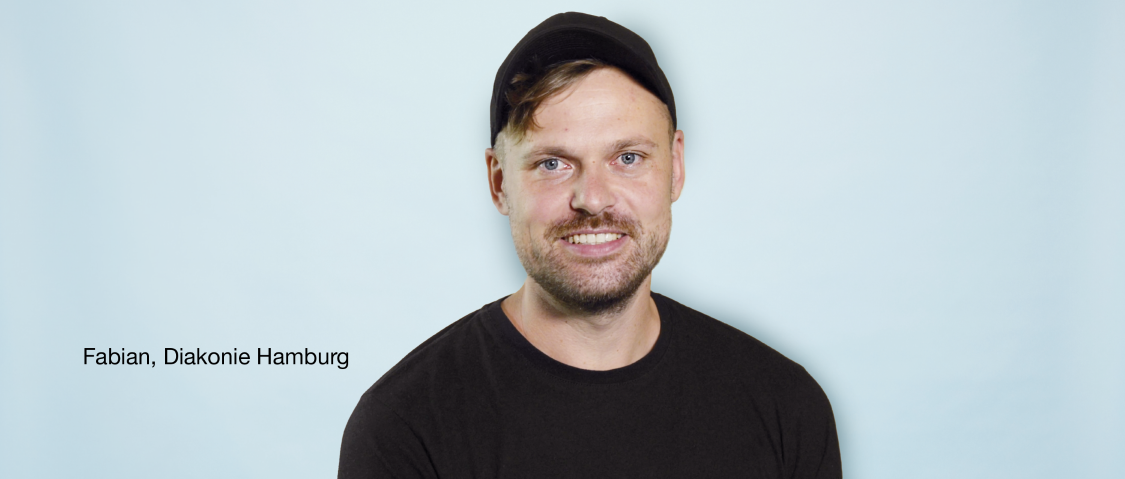 Person mit schwarzer Kappe und schwarzem T-Shirt lächelt vor hellblauem Hintergrund. Text links: „Fabian, Diakonie Hamburg“.