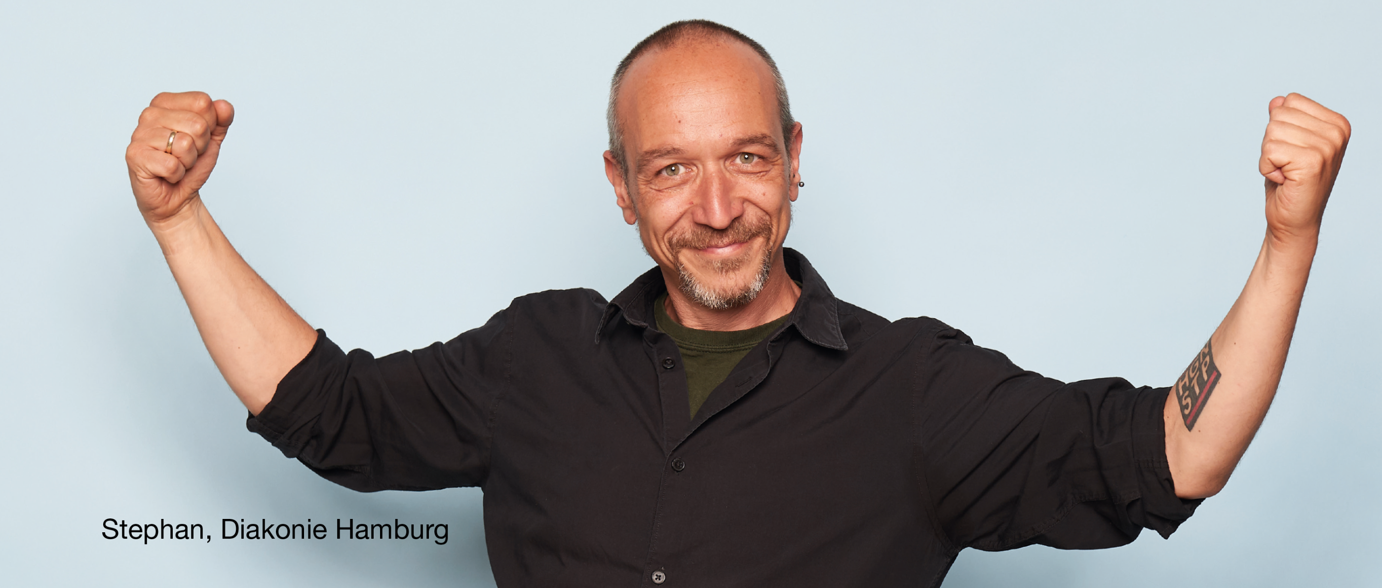 Person im schwarzen Hemd hebt beide Arme angewinkelt nach oben, Hände zu Fäusten geballt. Text im Bild: „Stephan, Diakonie Hamburg“.