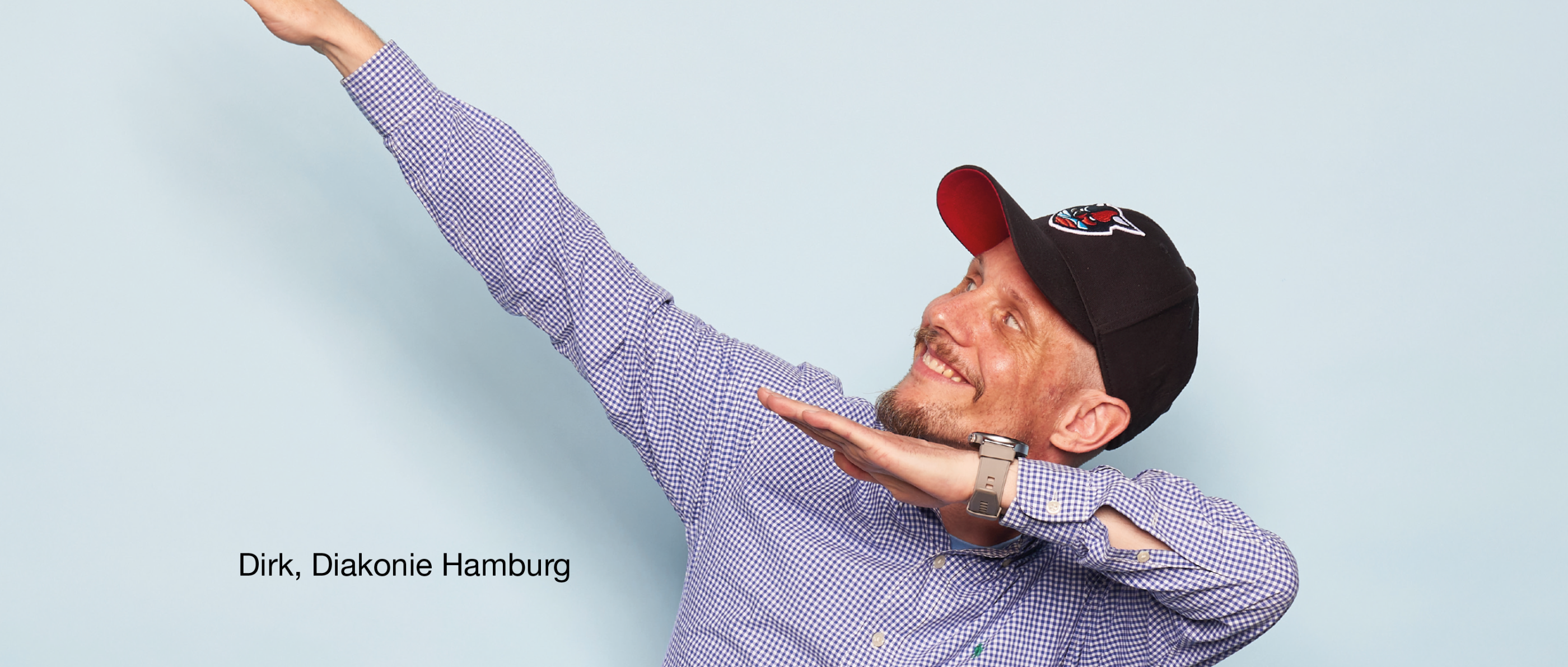 Person in kariertem Hemd und schwarzer Kappe posiert seitlich mit ausgestreckten Armen in dynamischer Haltung. Text im Bild: „Dirk, Diakonie Hamburg“.