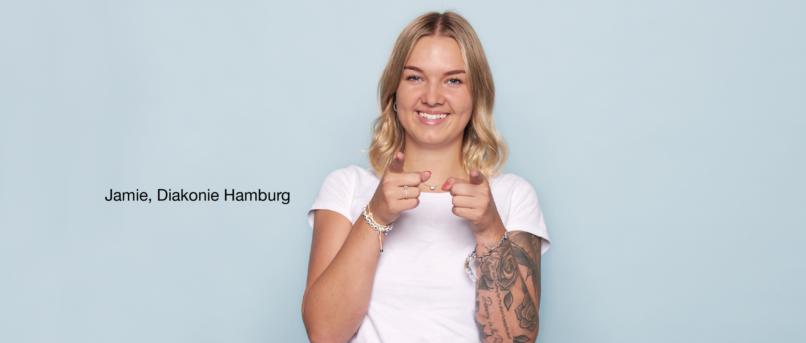 Person mit langen Haaren und sichtbaren Tattoos zeigt mit beiden Händen zur Kamera und lächelt. Text links: „Jamie, Diakonie Hamburg“.