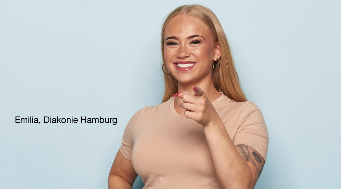 Person steht vor hellblauem Hintergrund, trägt ein beigefarbenes T-Shirt und zeigt mit einer Hand direkt nach vorne. Auf dem linken Arm ist ein Tattoo sichtbar. Links Schrift: 'Emilia, Diakonie Hamburg'.
