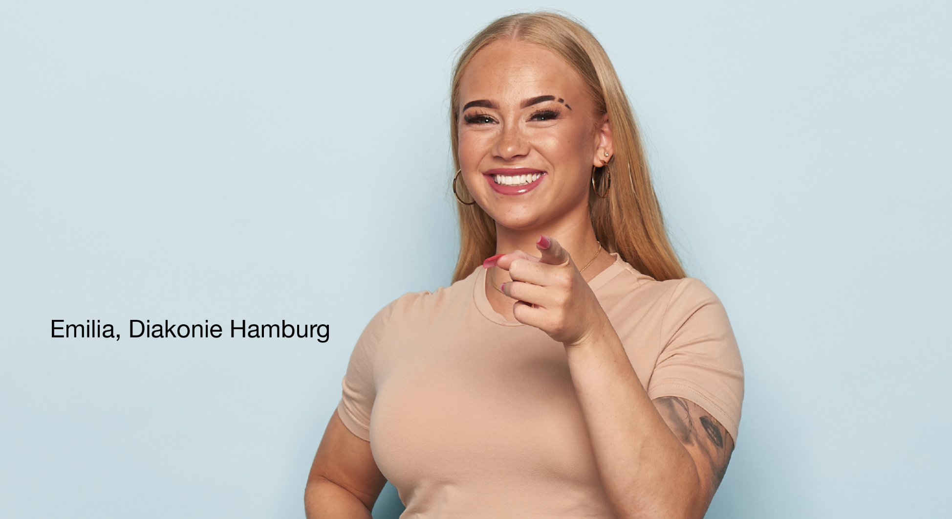 Person steht vor hellblauem Hintergrund, trägt ein beigefarbenes T-Shirt und zeigt mit einer Hand direkt nach vorne. Auf dem linken Arm ist ein Tattoo sichtbar. Links Schrift: 'Emilia, Diakonie Hamburg'.