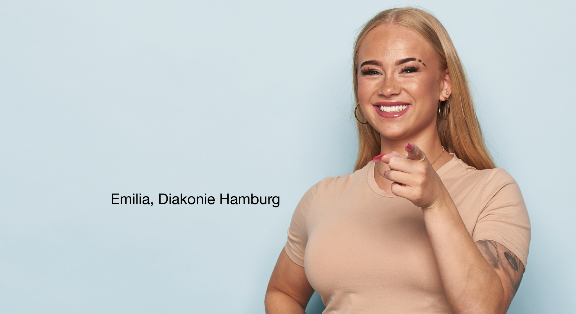 Person steht vor hellblauem Hintergrund, trägt ein beigefarbenes T-Shirt und zeigt mit einer Hand direkt nach vorne. Auf dem linken Arm ist ein Tattoo sichtbar. Links Schrift: 'Emilia, Diakonie Hamburg'.