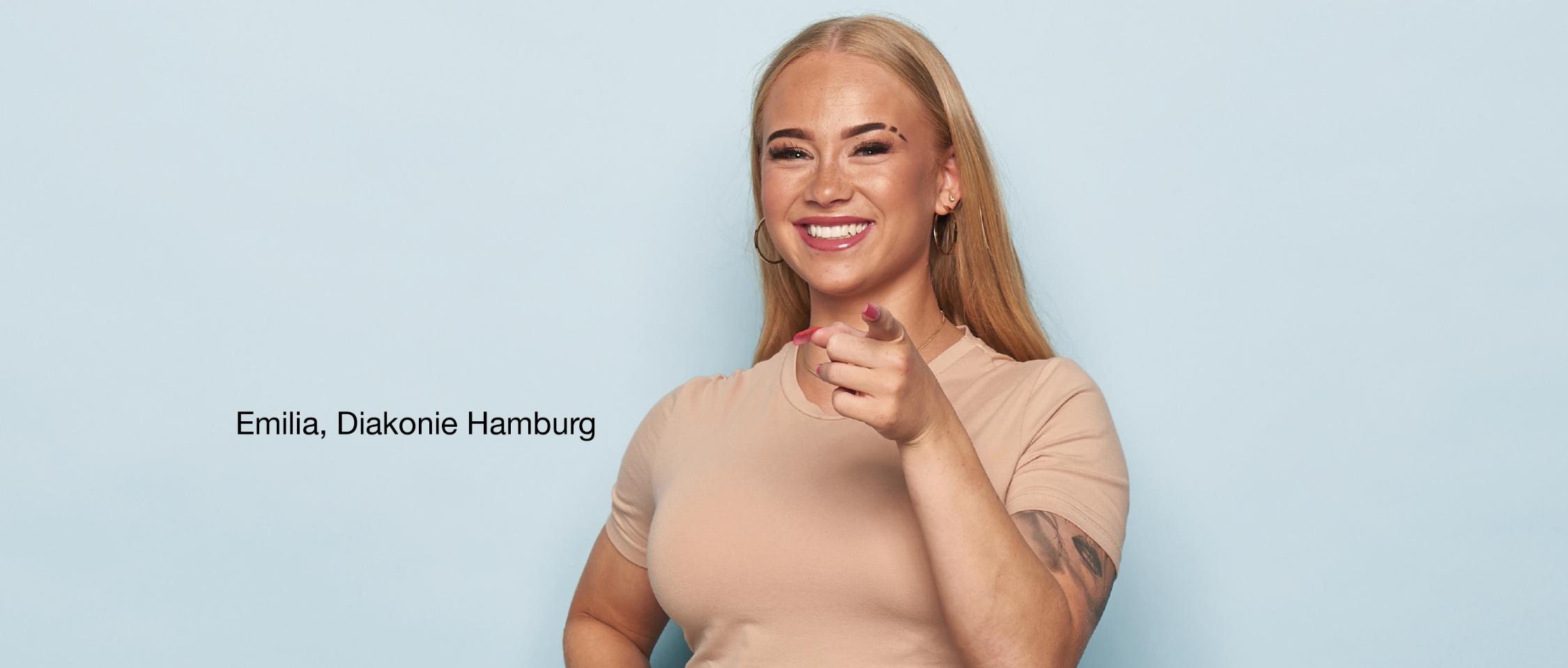 Person steht vor hellblauem Hintergrund, trägt ein beigefarbenes T-Shirt und zeigt mit einer Hand direkt nach vorne. Auf dem linken Arm ist ein Tattoo sichtbar. Links Schrift: 'Emilia, Diakonie Hamburg'.