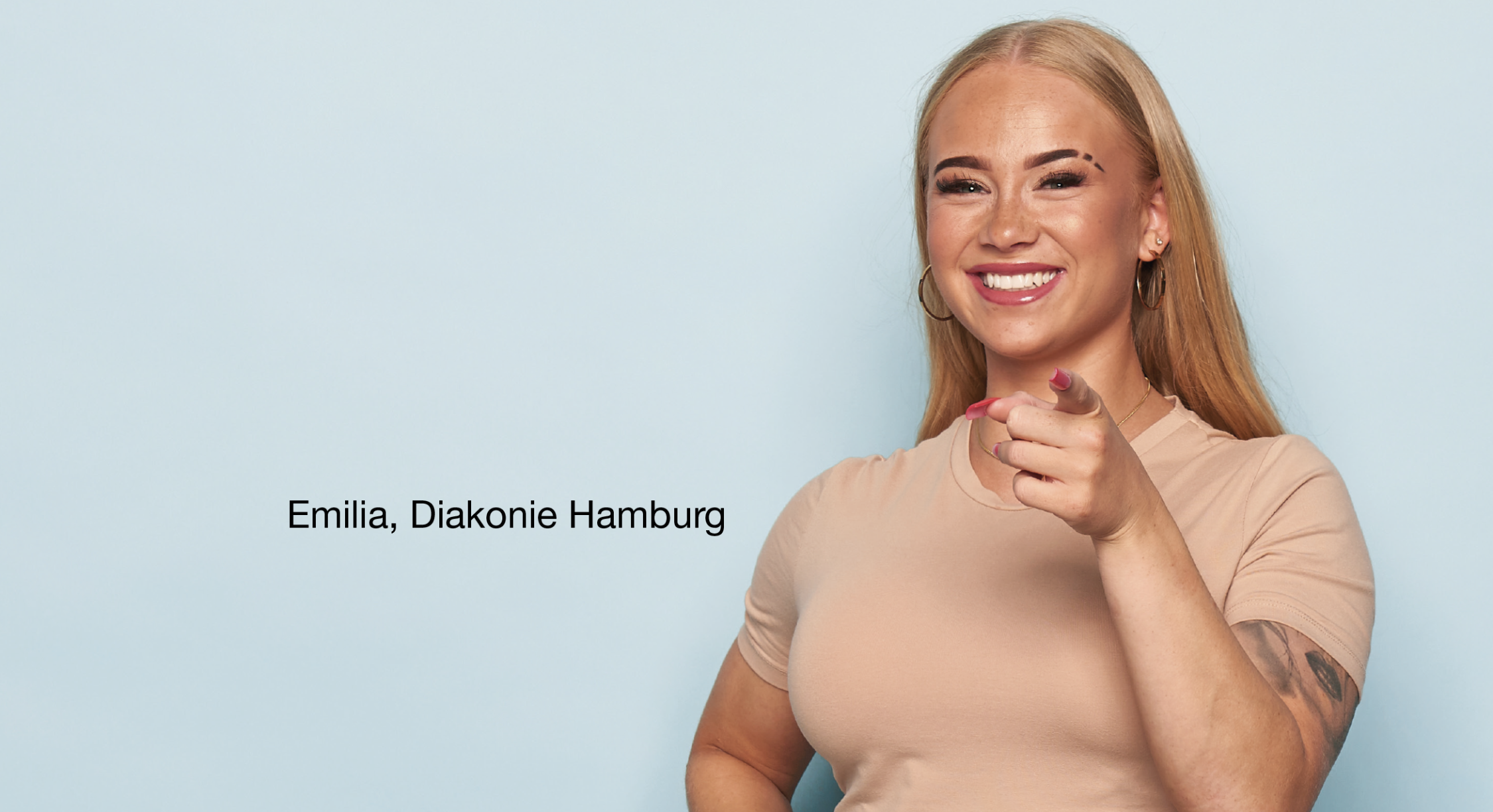 Person steht vor hellblauem Hintergrund, trägt ein beigefarbenes T-Shirt und zeigt mit einer Hand direkt nach vorne. Auf dem linken Arm ist ein Tattoo sichtbar. Links Schrift: 'Emilia, Diakonie Hamburg'.