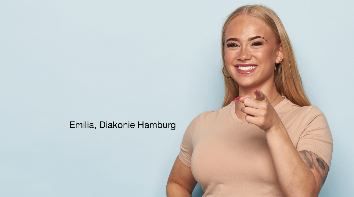 Person steht vor hellblauem Hintergrund, trägt ein beigefarbenes T-Shirt und zeigt mit einer Hand direkt nach vorne. Auf dem linken Arm ist ein Tattoo sichtbar. Links Schrift: 'Emilia, Diakonie Hamburg'.