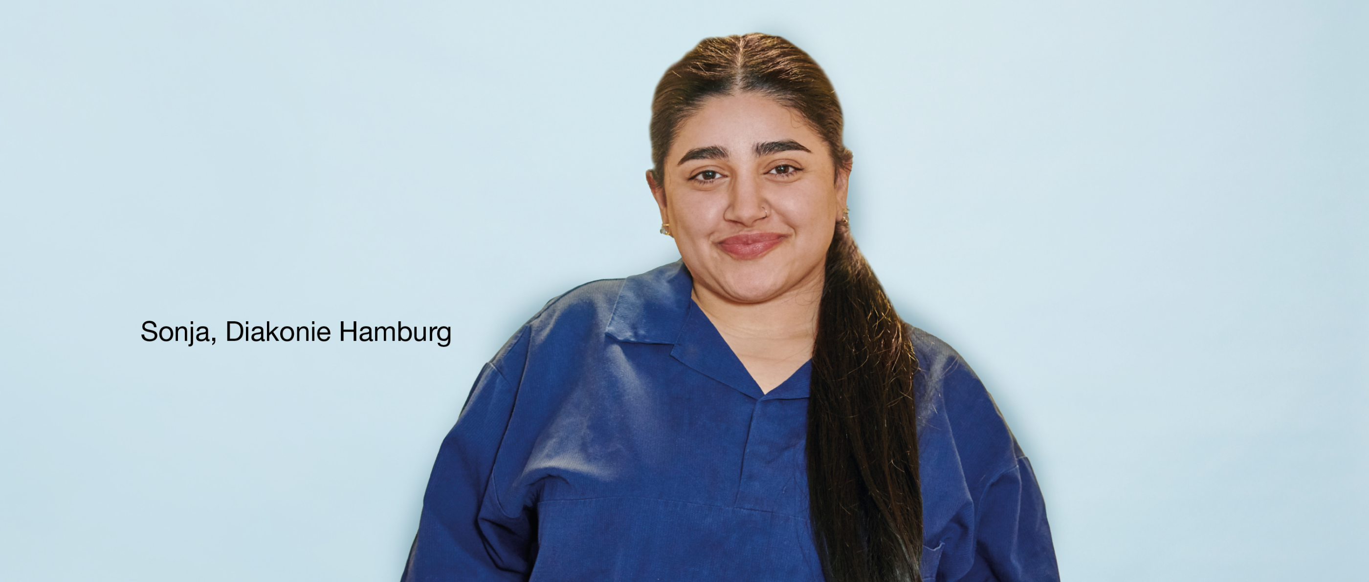Person mit zurückgebundenem dunklem Haar im blauen Arbeitskittel lächelt vor hellblauem Hintergrund. Text links: „Sonja, Diakonie Hamburg“.