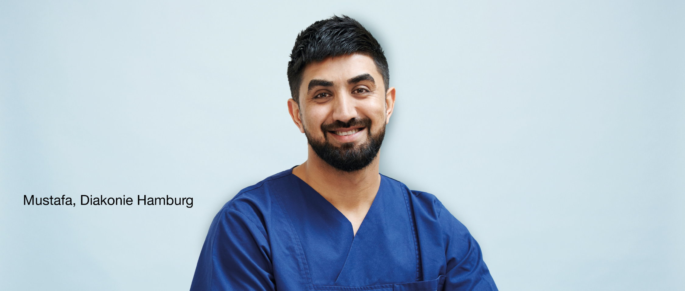 Person im blauen Arbeitskittel lächelt vor hellblauem Hintergrund. Text links: „Mustafa, Diakonie Hamburg“.