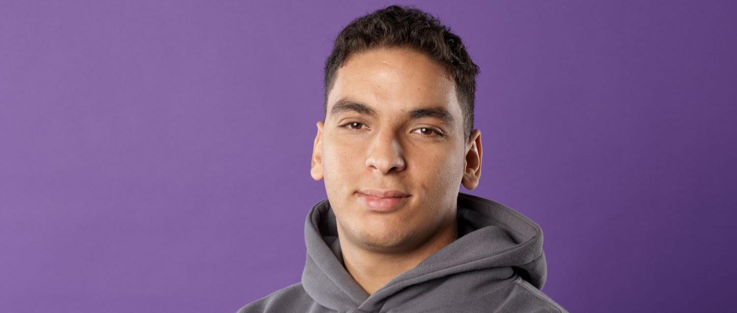 Person steht vor einfarbigem violettem Hintergrund, trägt einen grauen Kapuzenpullover und hat die Arme locker vor dem Körper verschränkt.