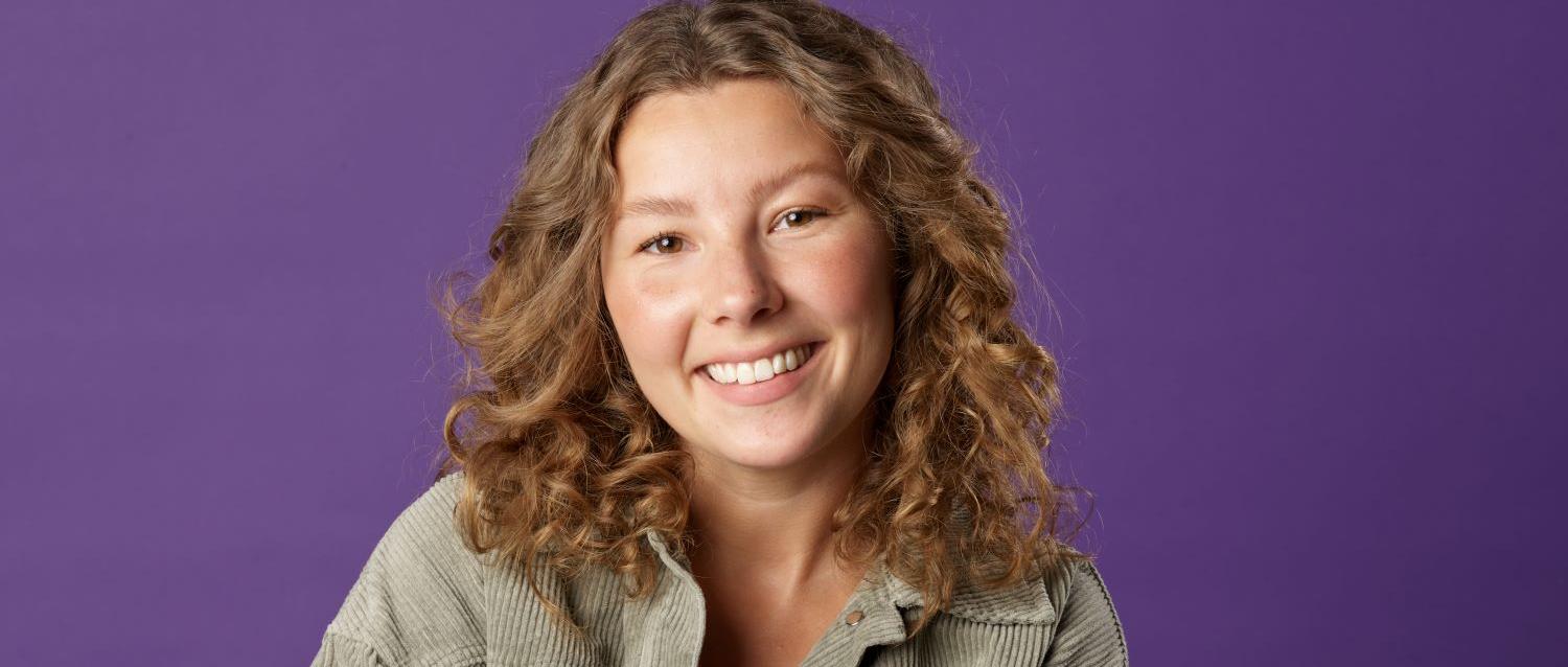 Person sitzt vor einfarbigem violettem Hintergrund, trägt ein orangefarbenes Oberteil unter einer offenen, grauen Cordjacke und hat die Hände locker vor dem Körper verschränkt.