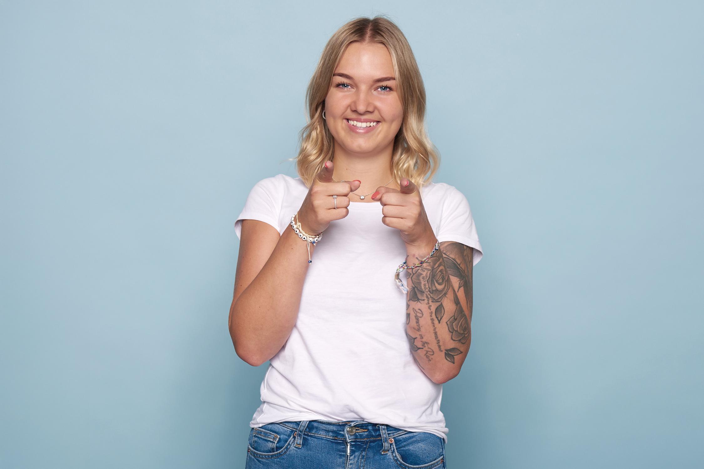 Person mit blonden Haaren und Tattoos zeigt selbstbewusst in die Kamera, vor hellblauem Hintergrund.